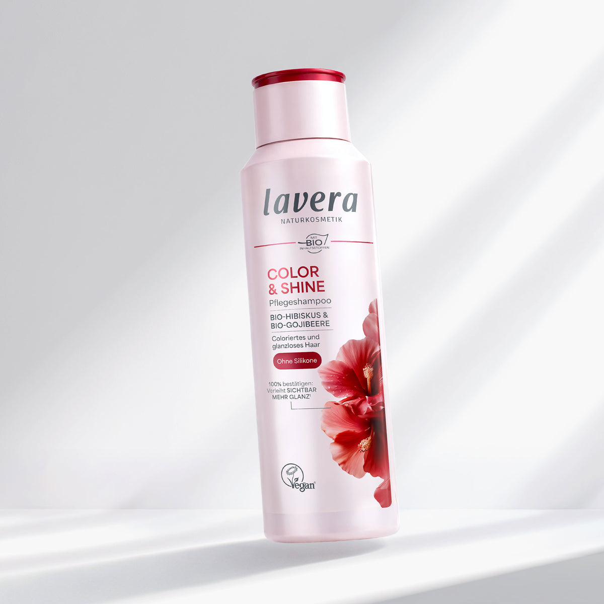 A bottle of Lavera Color & Shine hair shampoo, featuring a white bottle with a red cap and a red hibiscus flower graphic on the front, set against a clean white background with soft diagonal shadows.