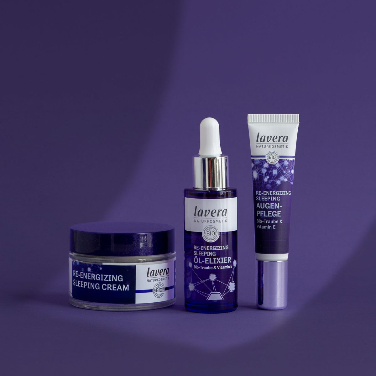 Three Lavera organic re-energizing sleeping skincare products arranged on a purple studio background: an re-energizing sleeping cream jar, a serum dropper bottle, and an eye-cream tube.