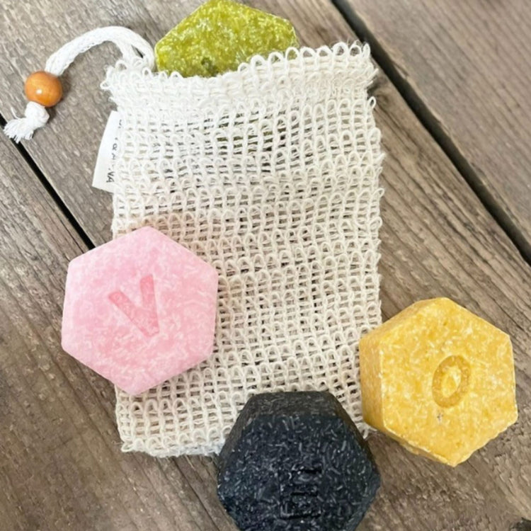 Four colourful Ben & Anna solid shampoo bars in hexagonal shapes (pink, yellow, green, and black) arranged on a wooden surface with a natural sisal soap saver bag.