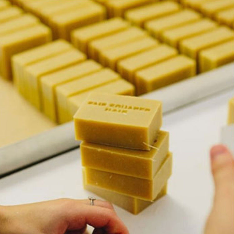 A stack of Fair Squared fair trade shampoo bars in a production setting. These rectangular, tan-colored solid soap bars are stamped with the brand name, emphasizing artisanal, plastic-free, and ethical manufacturing.
