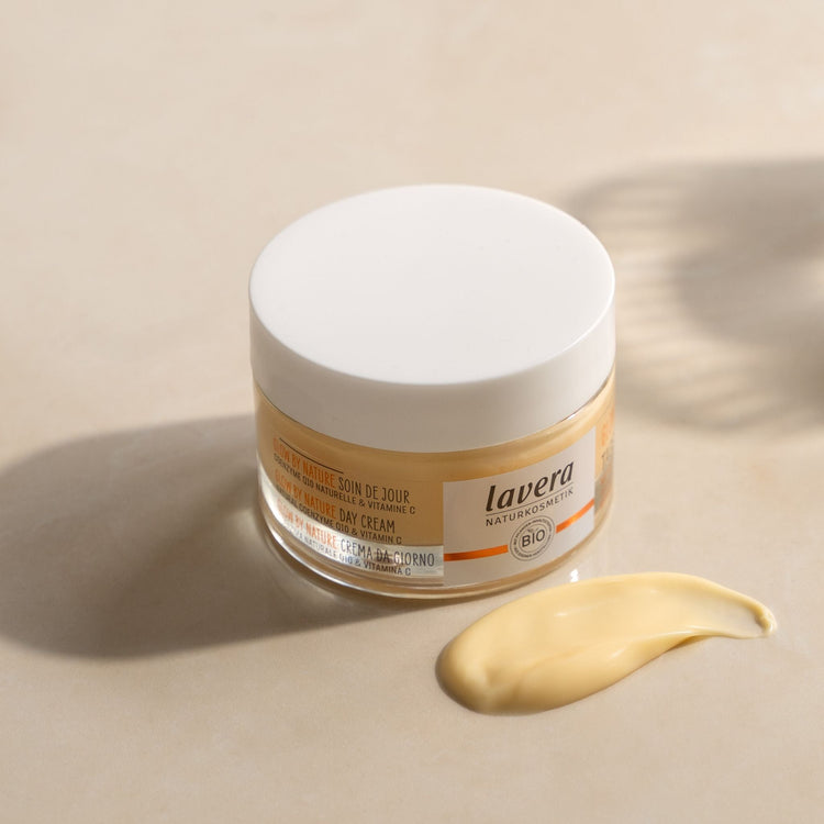 A clear glass jar of lavera Glow by Nature Day Cream with a white lid, featuring a smear of the smooth, pale yellow cream in the foreground. The label highlights key ingredients Natural Coenzyme Q10 and Vitamin C.