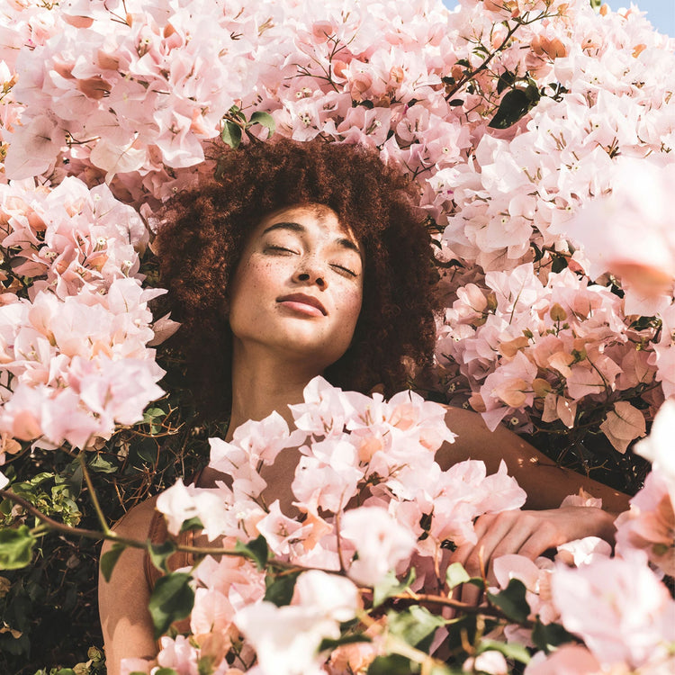 A woman with freckles and curly hair rests her head back with her eyes closed, surrounded by a dense bush of soft pink bougainvillea flowers. The lighting is warm and natural, creating a serene and gentle atmosphere.