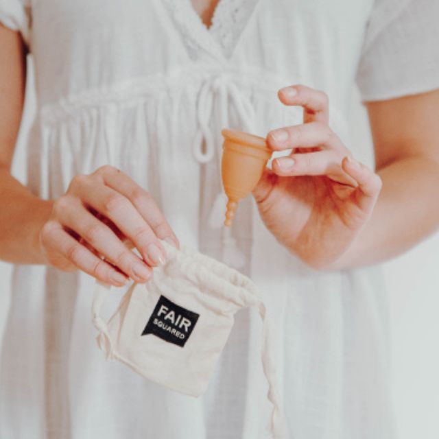 Person holding a menstrual cup and its packaging with 'FAIR SQUARED' branding.