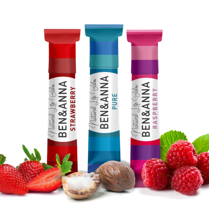 Three Ben & Anna lip balm tubes standing upright on a white background, surrounded by fresh strawberries, raspberries, and a nut. The tubes are labeled Natural Lip Balm in flavors Strawberry, Pure, and Raspberry.