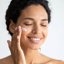 A close-up of a woman with closed eyes applying a dollop of white cream to her cheek with her finger against a clean white background.