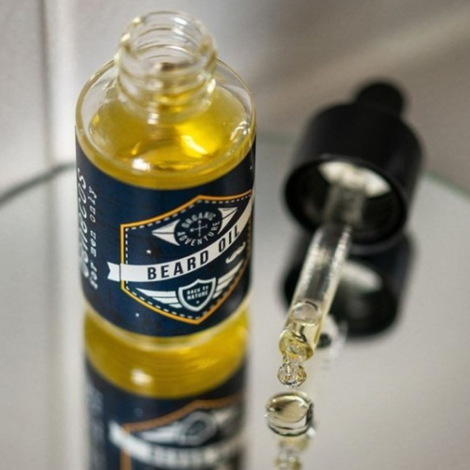 A clear glass bottle of benecos beard oil with a navy and gold label, shown next to an open black dropper bottle with oil droplets falling into a reflective surface.