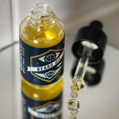 A clear glass bottle of benecos beard oil with a navy and gold label, shown next to an open black dropper bottle with oil droplets falling into a reflective surface.