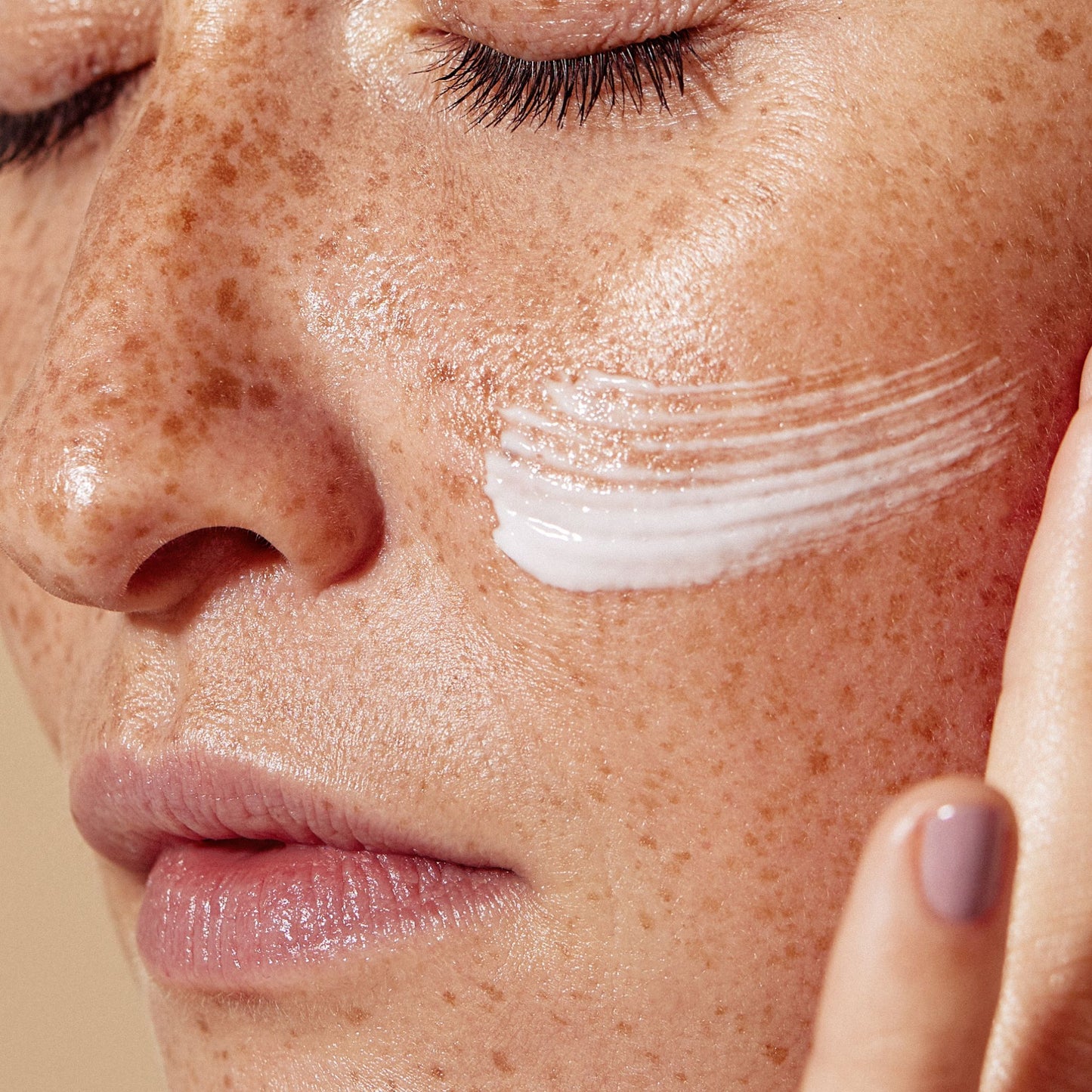 Close-up of a person's face with freckles, showing a swipe of white cream applied to the cheek and a finger touching the side of the face.