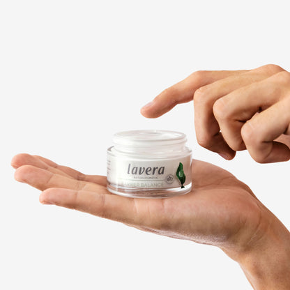 A small clear cosmetic jar labeled 'lavera Barrier Balance' resting on an open hand, with a person's fingers hovering above the lid against a plain light background.