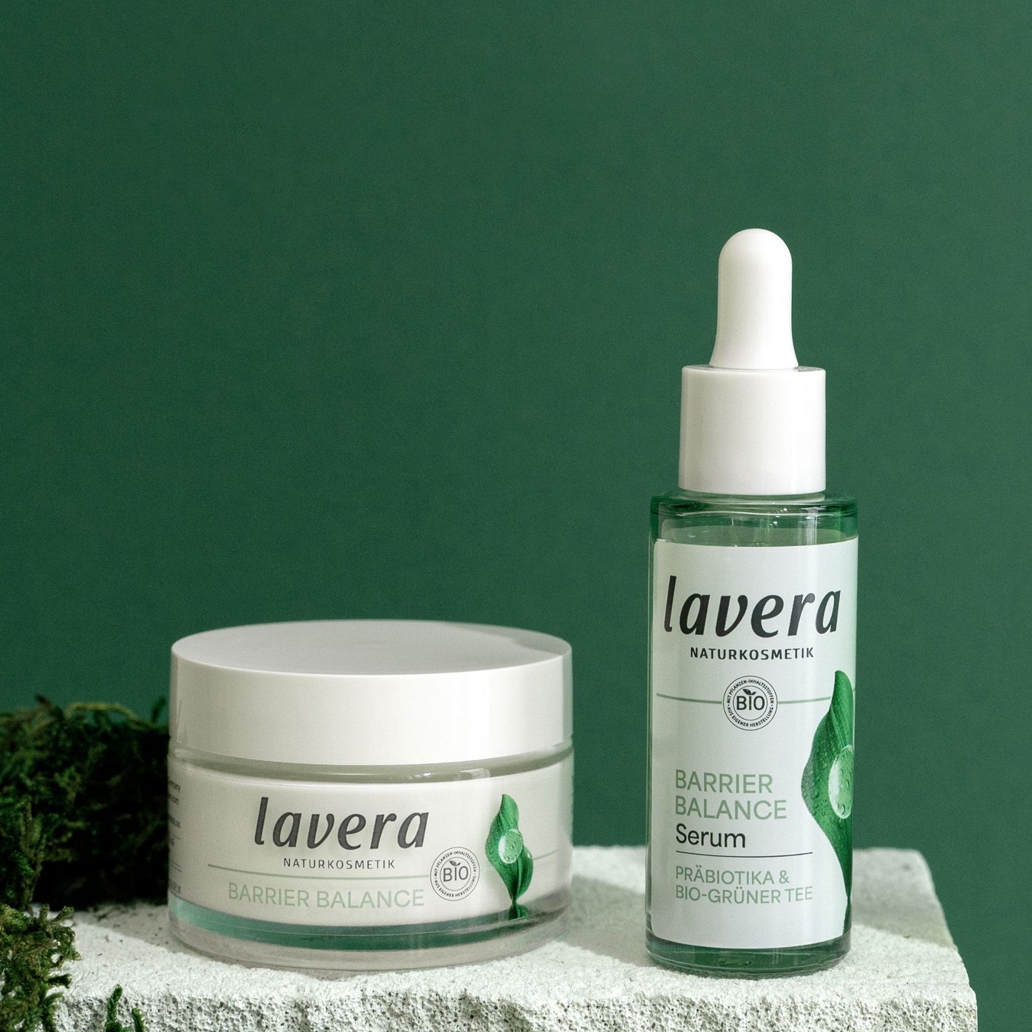 Lavera Barrier Balance skincare set featuring a clear serum bottle with a white dropper cap and a white cream jar, displayed on a textured stone surface against a dark green background.