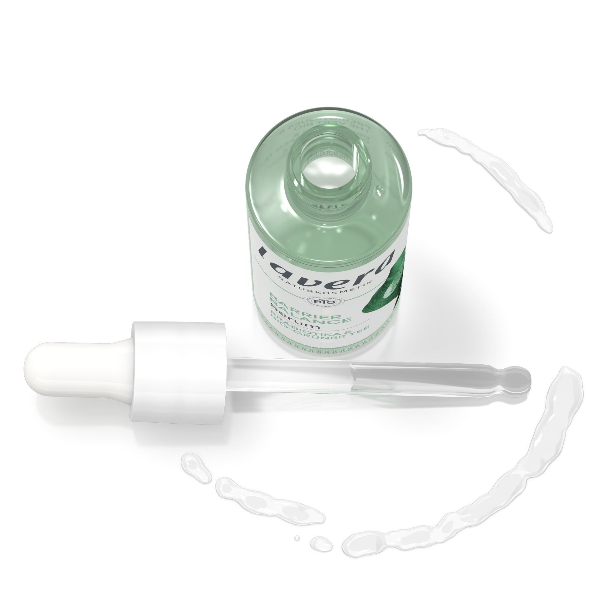 A clear glass dropper bottle with a white dropper and a pale mint green liquid inside, shown on a white background with a few droplets of the liquid scattered around it. The label shows the brand name lavera.