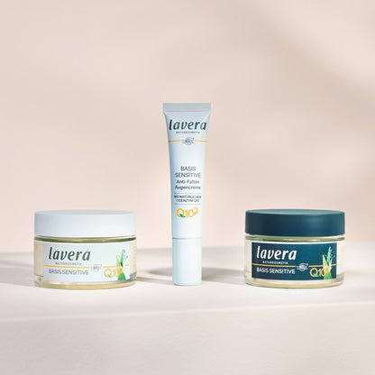 Three Lavera skincare products arranged on a light beige surface against a soft beige background: a clear jar with a white lid on the left, a white tube in the center, and a clear jar with a dark teal lid on the right. The products are labeled Basis Sensitive and Q10.