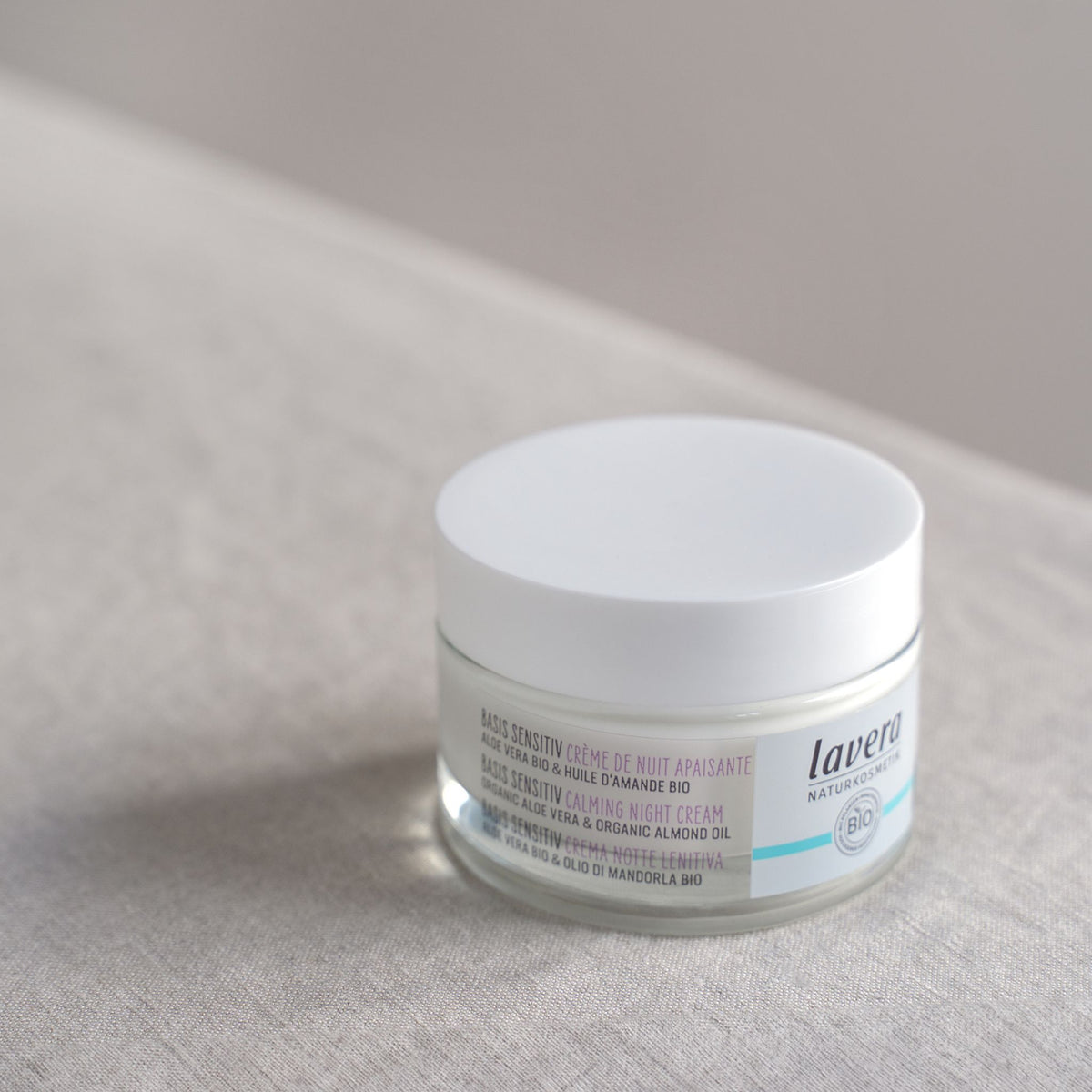 A jar of Lavera 'Basis Sensitive Calming Night Cream' on a textured background. The product is in a clear jar showing the white cream inside, with a white lid and a white label featuring purple text.