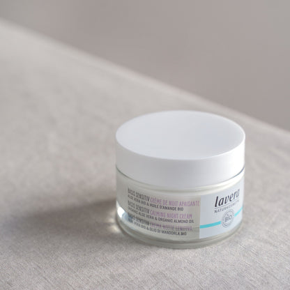A jar of Lavera 'Basis Sensitive Calming Night Cream' on a textured background. The product is in a clear jar showing the white cream inside, with a white lid and a white label featuring purple text.