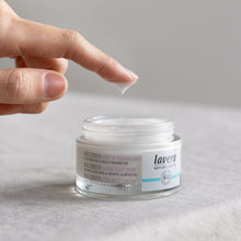 A jar of Lavera 'Basis Sensitive Calming Night Cream' An open round cosmetic jar on a textured background. The product is in a clear jar showing the white cream inside, with a white lid and a white label featuring purple text.