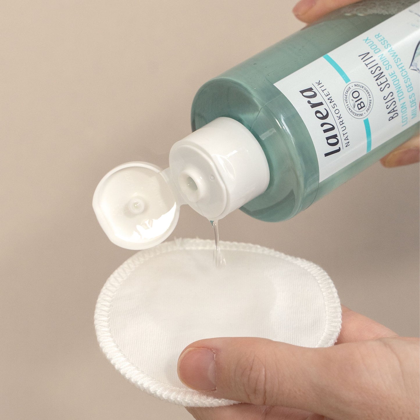 A hand holds a clear bottle of Lavera Basic Sensitive toner, pouring clear liquid onto a white makeup remover pad against a neutral beige background.