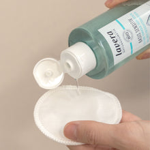 A hand holds a clear bottle of Lavera Basic Sensitive toner, pouring clear liquid onto a white makeup remover pad against a neutral beige background.