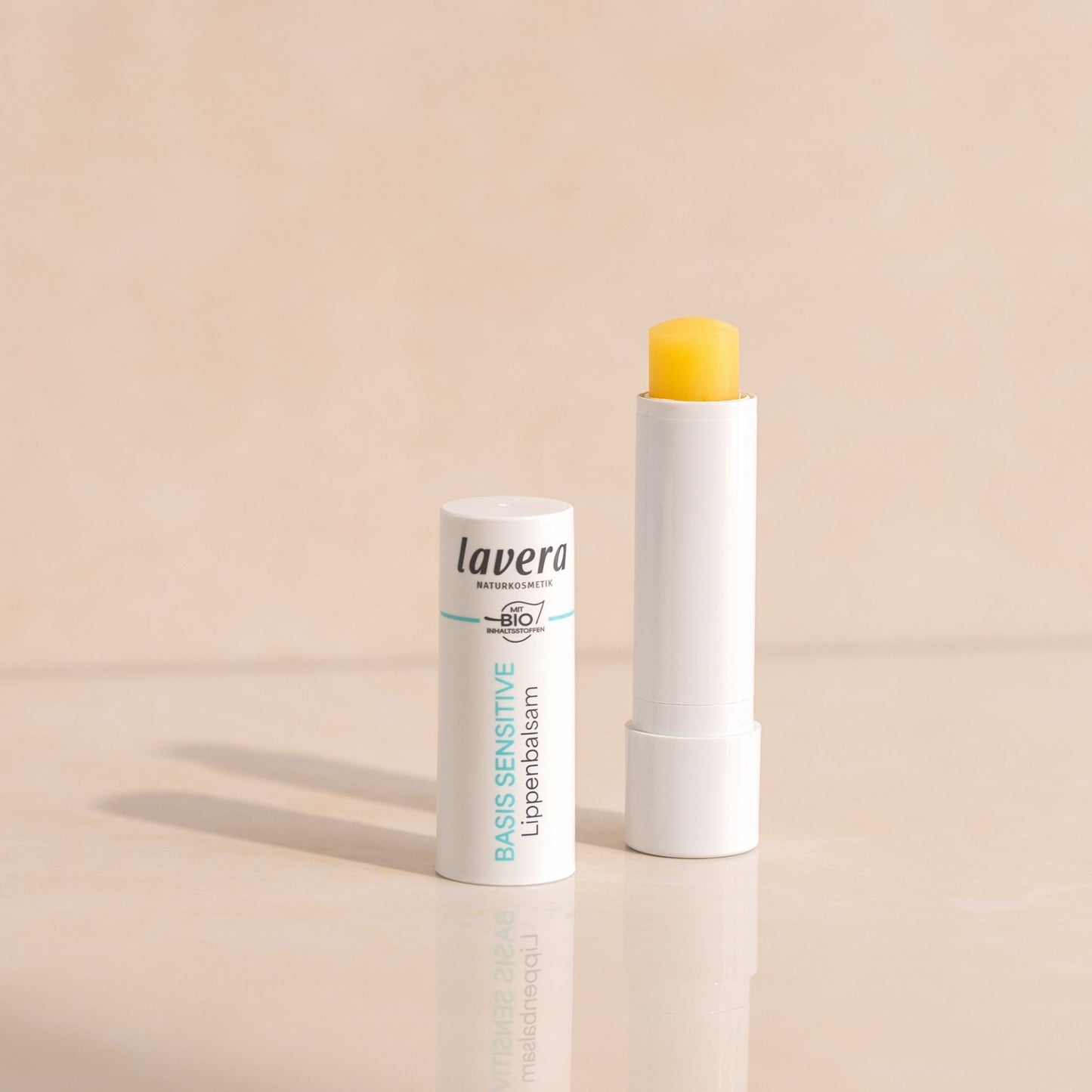 Lavera Basic Sensitive Lip Balm shown in its white tube packaging, with open cap standing upright and another tube open to reveal a bright yellow balm stick, set against a warm beige background.