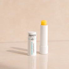 Lavera Basic Sensitive Lip Balm shown in its white tube packaging, with open cap standing upright and another tube open to reveal a bright yellow balm stick, set against a warm beige background.