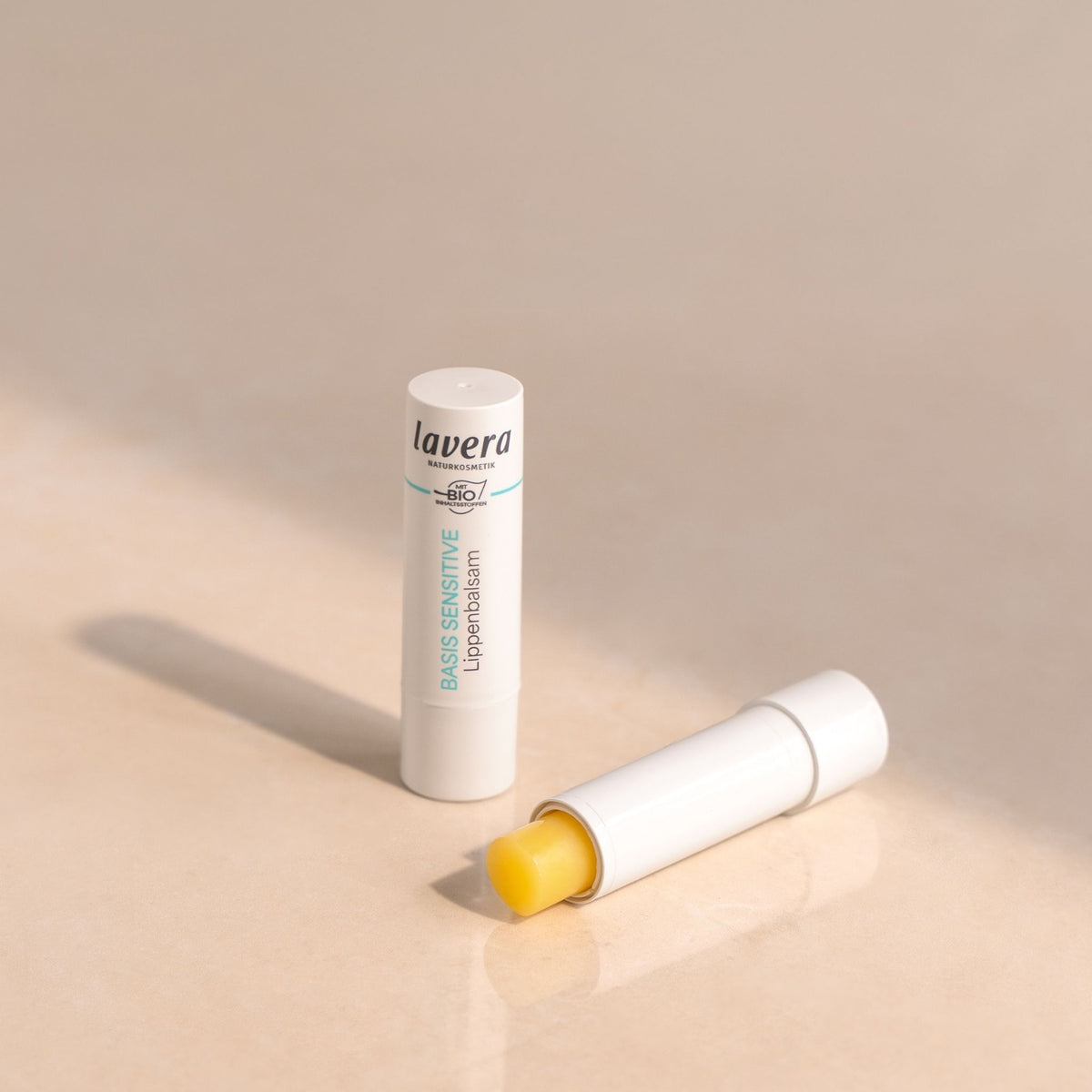 Lavera Basis Sensitive Lip balm shown in its white tube packaging, with one tube standing upright and another lying on its side to reveal the yellow balm stick inside, set against a warm beige background.