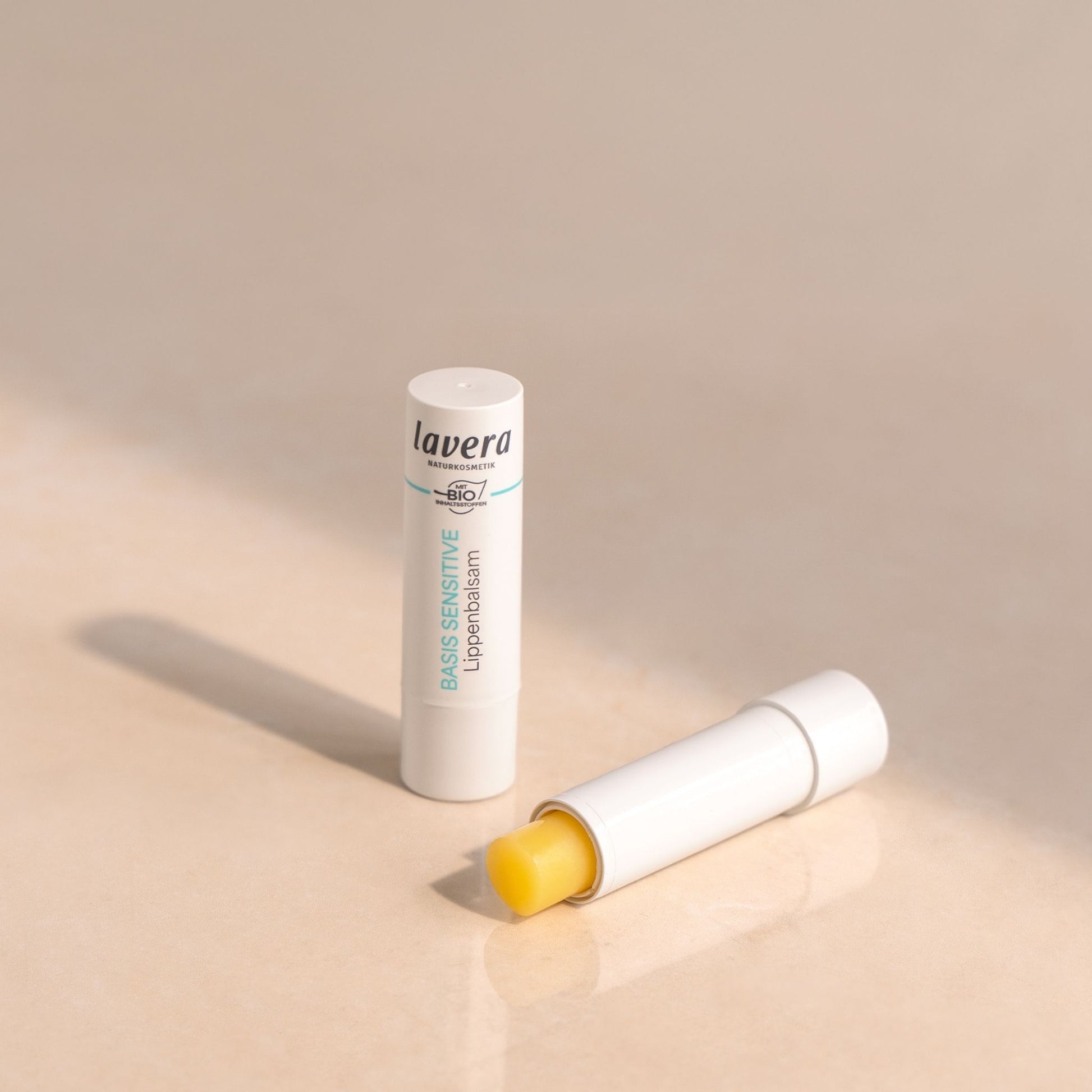 Lavera Basis Sensitive Lip balm shown in its white tube packaging, with one tube standing upright and another lying on its side to reveal the yellow balm stick inside, set against a warm beige background.