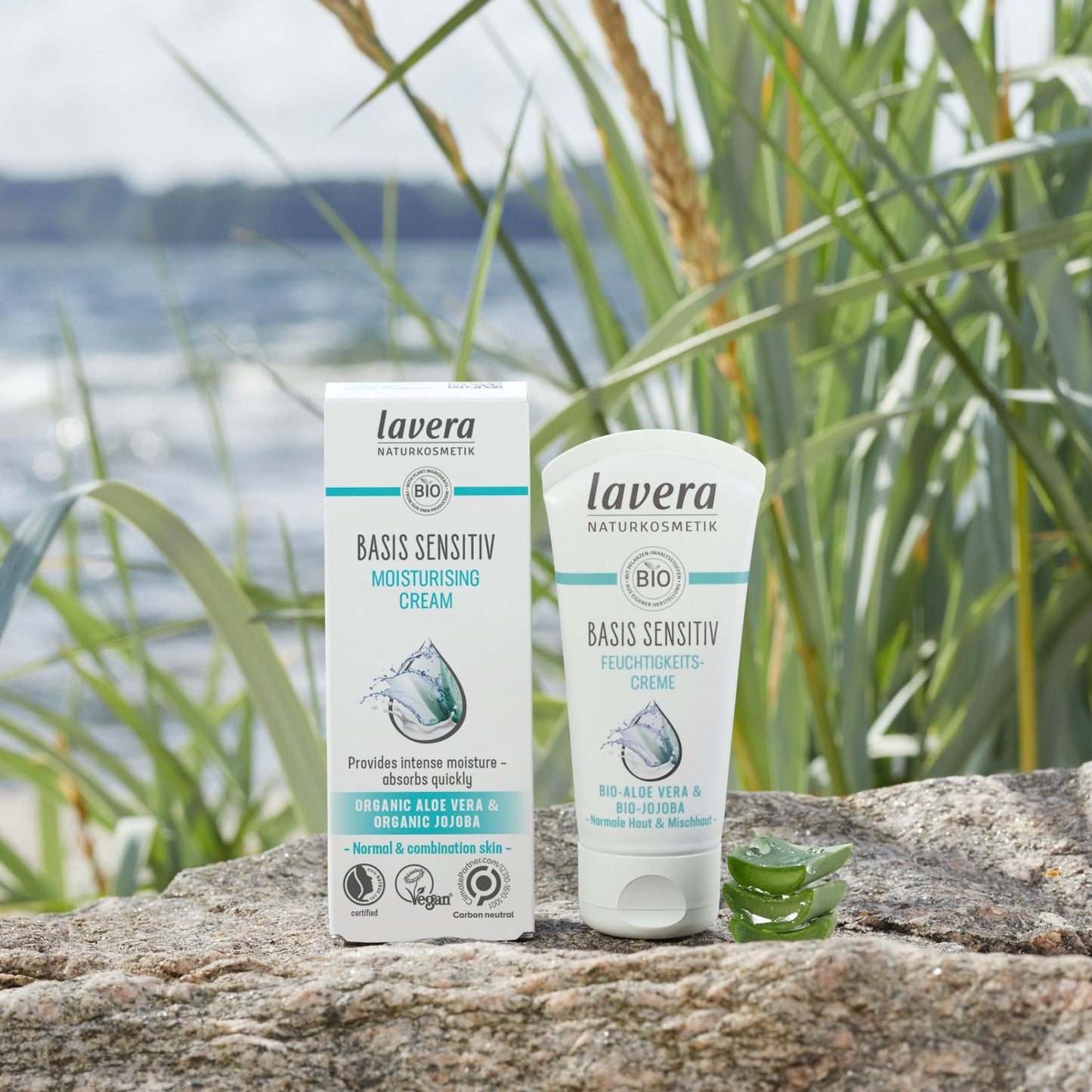 Lavera Basis Sensitive Moisturising Cream shown in its white and teal packaging box and squeeze tube, placed on a stone surface outdoors with reeds and water in the background.