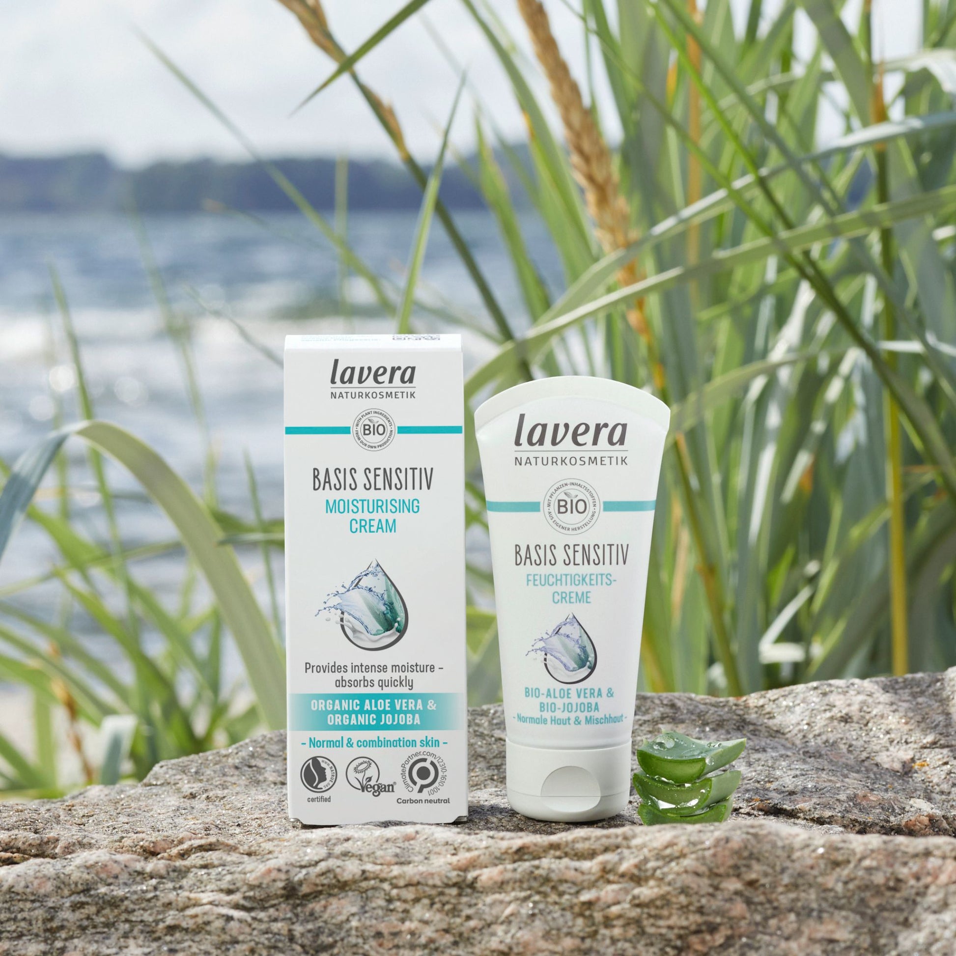Lavera Basis Sensitive Moisturising Cream shown in its white and teal packaging box and squeeze tube, placed on a stone surface outdoors with reeds and water in the background.