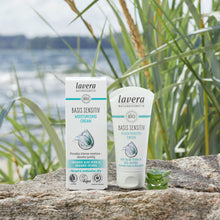 Lavera Basis Sensitive Moisturising Cream shown in its white and teal packaging box and squeeze tube, placed on a stone surface outdoors with reeds and water in the background.
