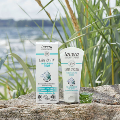 Lavera Basis Sensitive Moisturising Cream shown in its white and teal packaging box and squeeze tube, placed on a stone surface outdoors with reeds and water in the background.
