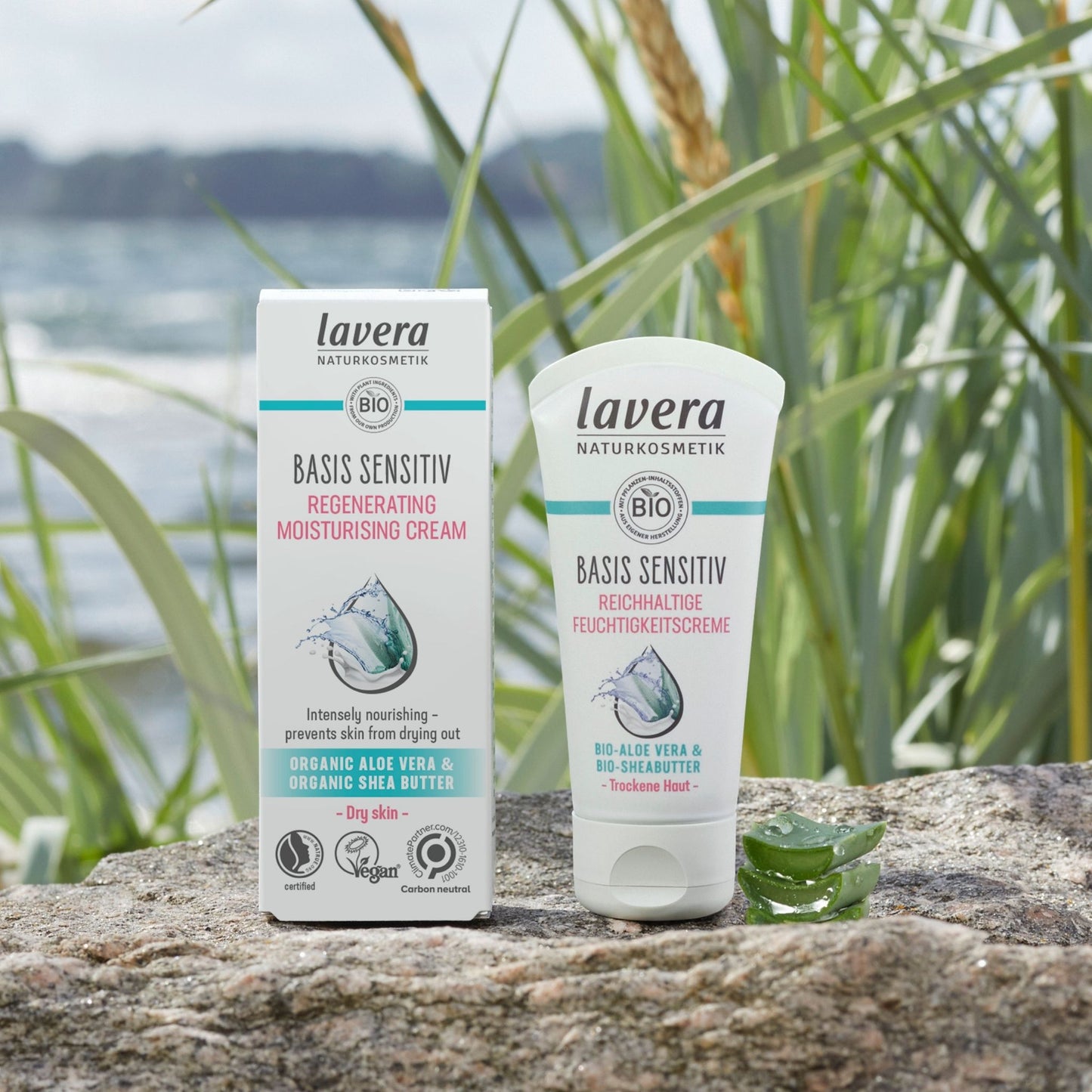 Lavera Basis Sensitive Regenerating Moisturising Cream shown as a white box and matching squeeze tube standing on a stone surface outdoors, with reeds and water blurred in the background.