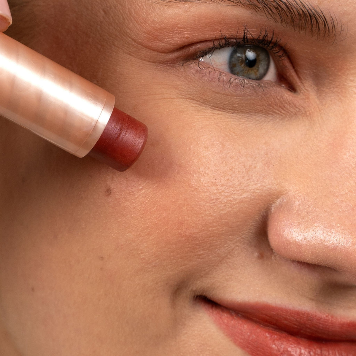Close-up of a person's face with a makeup stick being applied to the cheek. The product tube is metallic rose gold, and the stick itself is a warm reddish-brown color.