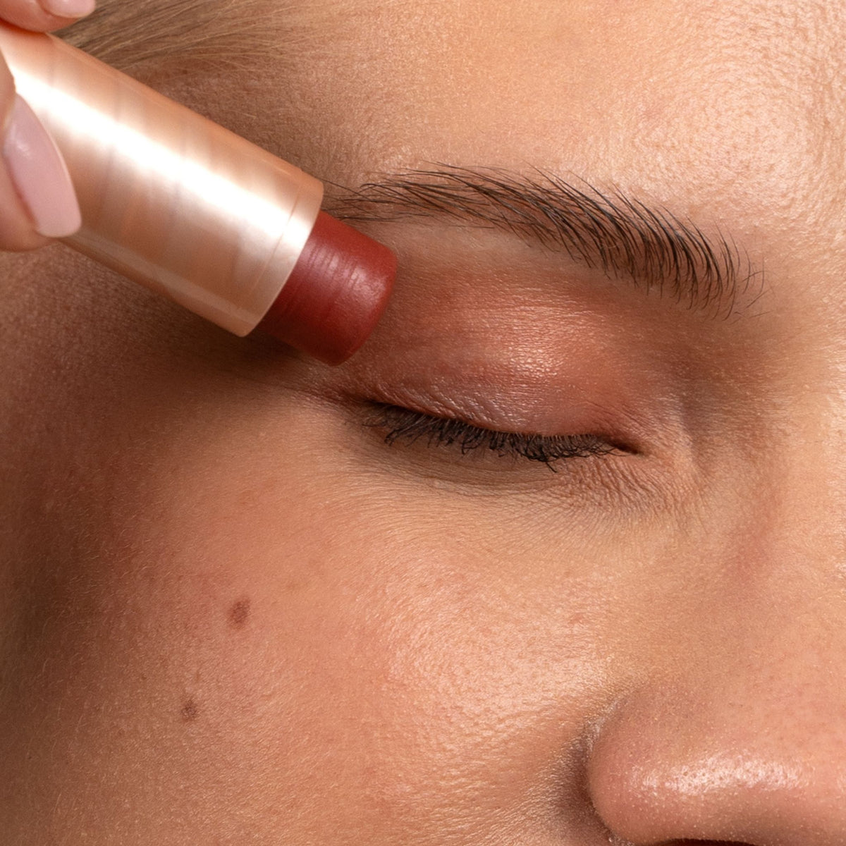 Close-up of a person's face with closed eyes as a makeup stick is being applied to the eyebrow with a metallic rose gold tube.