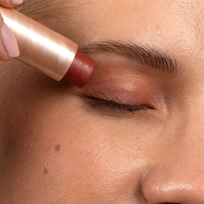 Close-up of a person's face with closed eyes as a makeup stick is being applied to the eyebrow with a metallic rose gold tube.