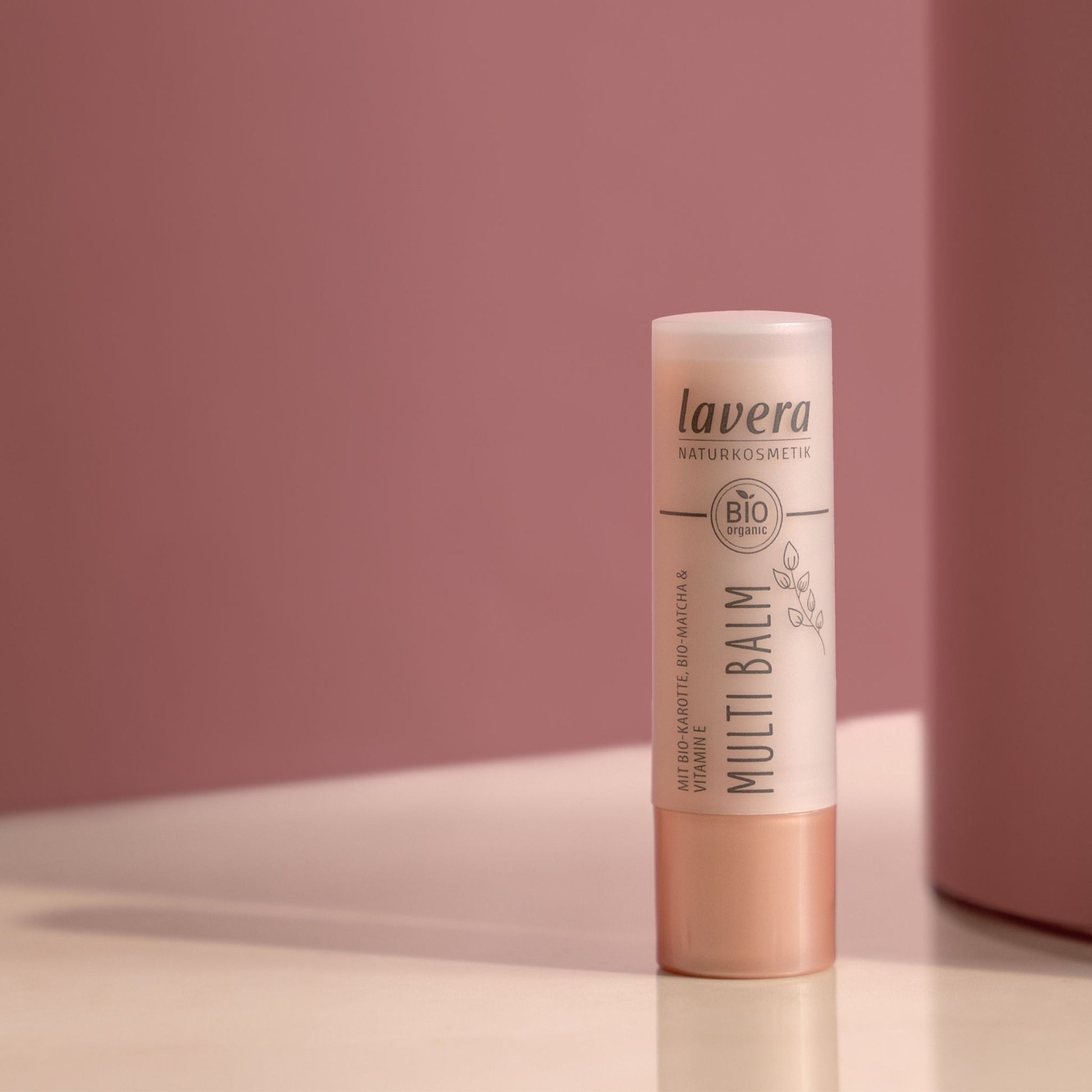 A single stick of Lavera Bio Certified Multi Balm lip balm standing upright on a white surface against a dusty pink background.