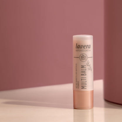A single stick of Lavera Bio Certified Multi Balm lip balm standing upright on a white surface against a dusty pink background.