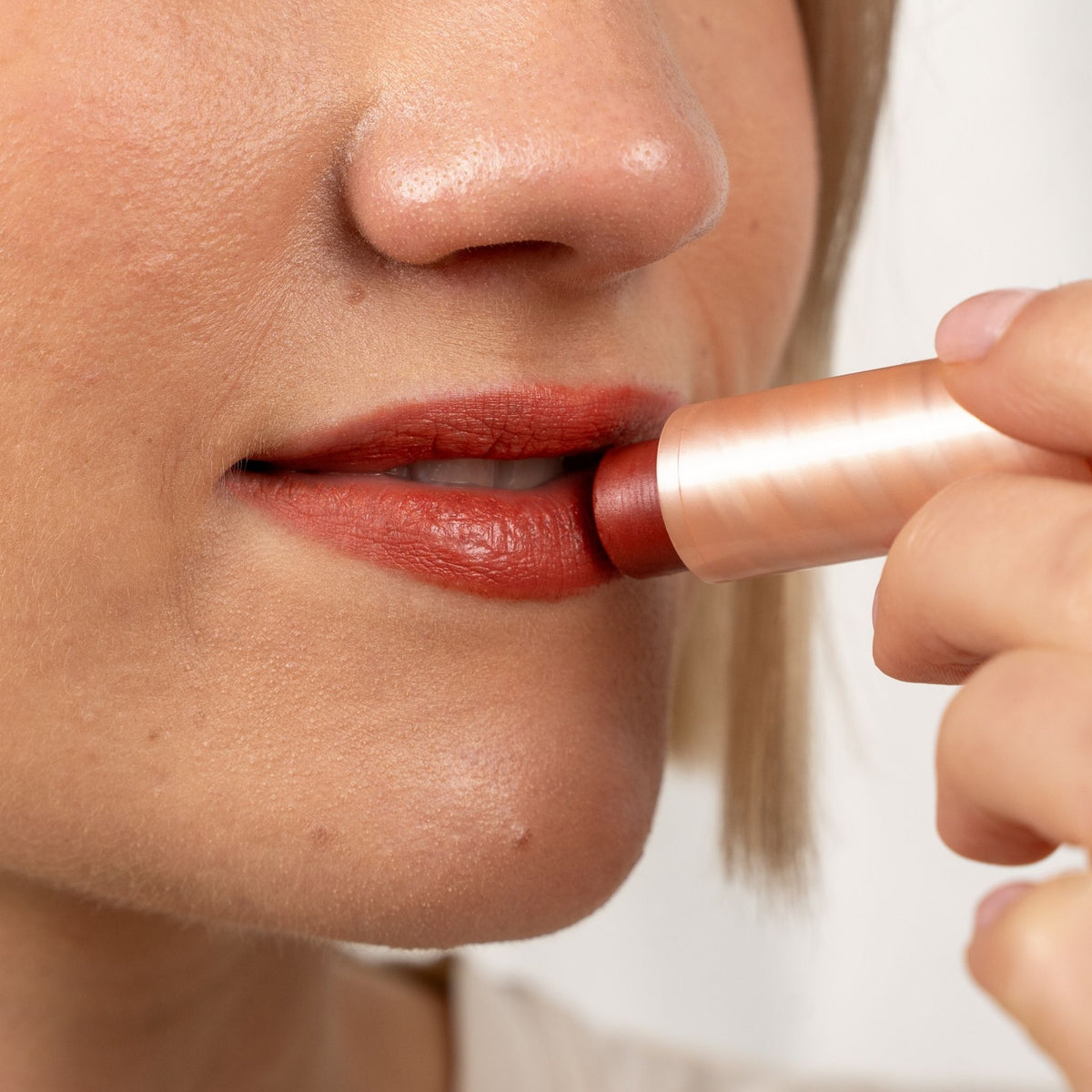 Close-up of a person applying a warm reddish-brown lipstick from a metallic rose gold tube.