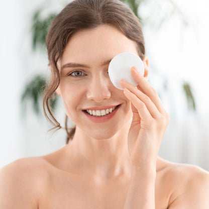 A smiling woman with her hair pulled back holds a white round makeup sponge to her eye, set against a bright, softly blurred indoor background with hints of greenery.