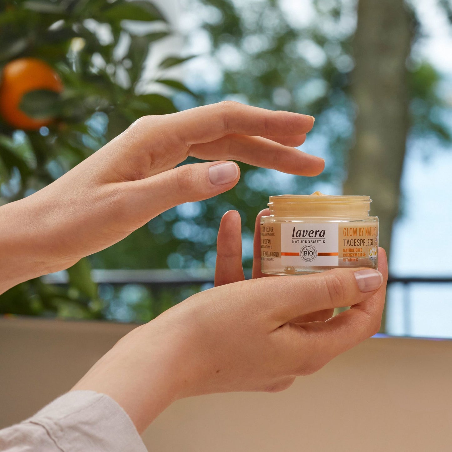 A person holds a small jar of Lavera Glow by Nature Day Cream, with their other hand hovering near the open lid. The background is a soft-focus outdoor scene with green foliage and a tree.