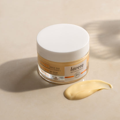 A jar of Lavera Glow by Nature Day Cream with a white lid and clear glass container, shown next to a smear of the pale yellow cream on a beige background.