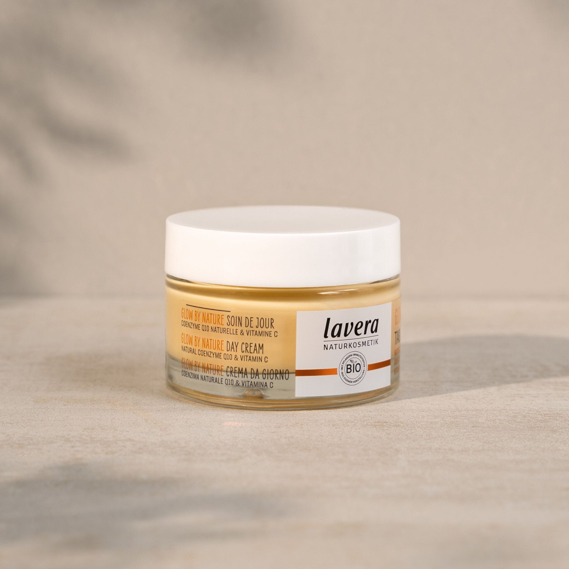 A jar of Lavera Glow by Nature Day Cream on a neutral beige textured surface with a soft, blurred background.