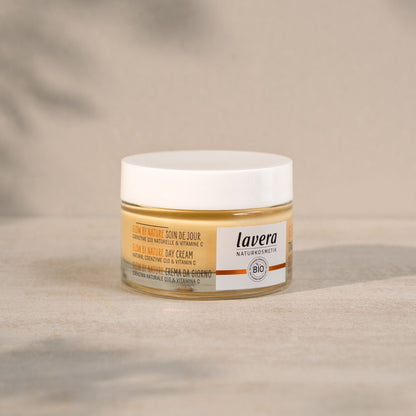 A jar of Lavera Glow by Nature Day Cream on a neutral beige textured surface with a soft, blurred background.