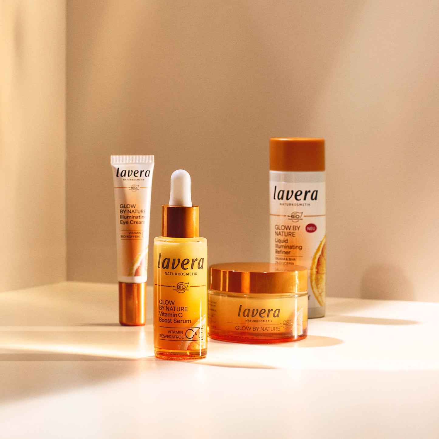 Four Lavera Glow by Nature facecare products arranged on a white surface with warm sunlight and shadows: an eye cream tube, a serum dropper bottle, a face cream, and a liquid refiner bottle, all in amber and white packaging.