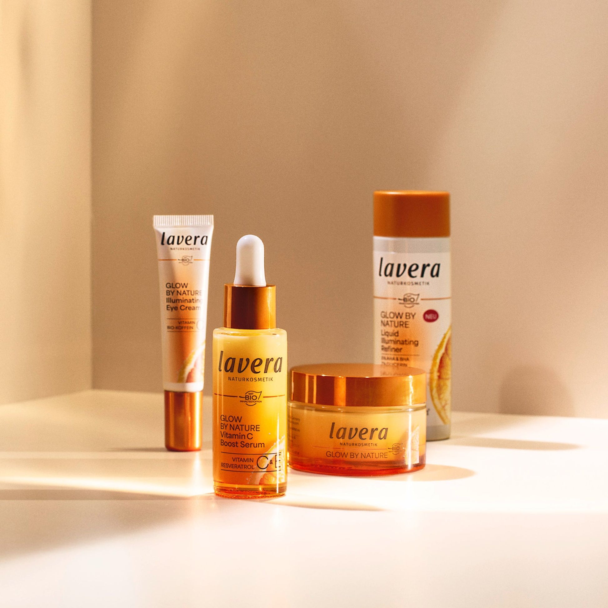 Four Lavera Glow by Nature facecare products arranged on a white surface with warm sunlight and shadows: an eye cream tube, a serum dropper bottle, a face cream, and a liquid refiner bottle, all in amber and white packaging.