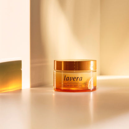 A jar of Lavera Glow by Nature cream with amber-colored gel and a metallic gold lid, set on a warm beige surface with dramatic side lighting.