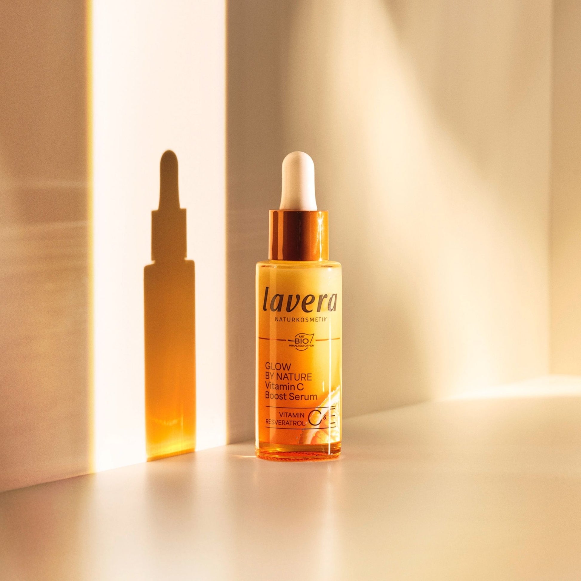 Lavera Glow by Nature Vitamin C Boost Serum in an amber glass dropper bottle with a white cap, shown next to its matching amber dropper bottle silhouette on a warm beige background.