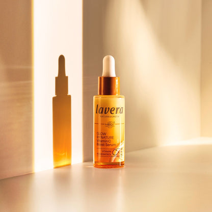 Lavera Glow by Nature Vitamin C Boost Serum in an amber glass dropper bottle with a white cap, shown next to its matching amber dropper bottle silhouette on a warm beige background.