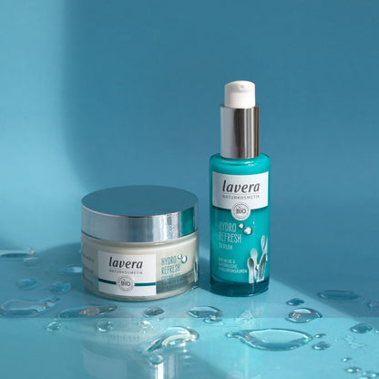 Two Lavera Hydro Refresh skincare products on a glossy blue surface with water droplets: a white and silver jar labeled Hydra Refresh Cream-Gel and a turquoise pump bottle labeled Organic Hydra Refresh Serum.