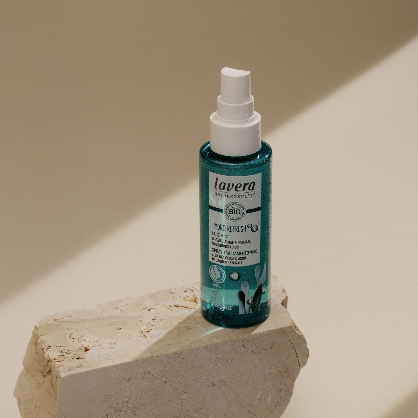 A bottle of Lavera Bio Refresher face mist, standing on a textured stone pedestal against a warm beige background with soft diagonal shadows.