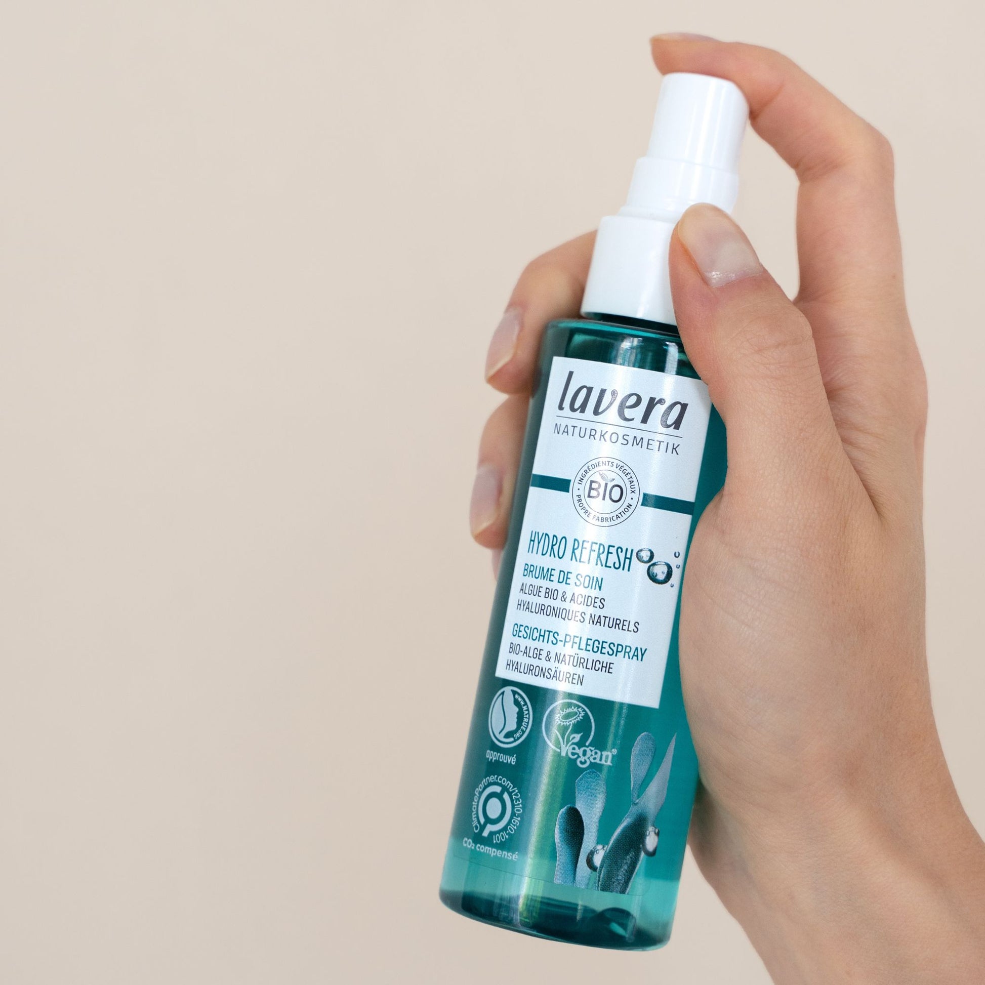 A hand holding a bottle of Lavera Bio Hydro Refresh Face Mist Spray against a solid beige background.