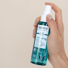 A hand holding a bottle of Lavera Bio Hydro Refresh Face Mist Spray against a solid beige background.
